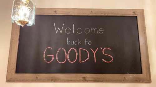 Chalkboard sign reading "Welcome back to GOODY'S" framed in wood, with a decorative light fixture above.