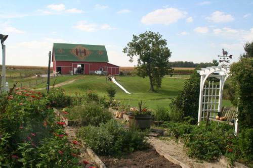 A red barn with a green roof sits in a lush landscape, featuring a slide, trees, and colorful flowers in the foreground.