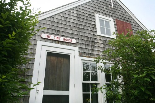 A small, rustic house with wooden shingles and a sign reading "The Tiny House," surrounded by greenery.