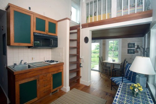Cozy kitchen with wooden cabinets, a staircase to a loft, and a small dining area with a view of greenery outside.