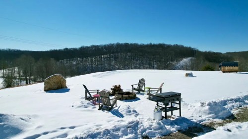 A snowy landscape with a fire pit surrounded by chairs, set against a backdrop of rolling hills and clear blue sky.