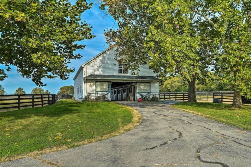 A white barn with a gravel driveway, surrounded by green grass and trees under a blue sky.