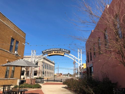 Archway sign for Tanglefoot Trail in a small town, with buildings and a clear blue sky in the background.