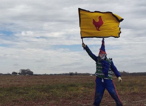 A scarecrow in a colorful outfit holds a yellow flag with a red chicken silhouette against a cloudy sky.