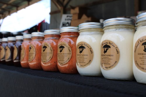 A row of jars filled with various sauces, each labeled, displayed on a black tablecloth at a market.