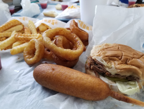 A close-up of a meal featuring a burger, corndog, onion rings, and crinkle-cut fries on a table.