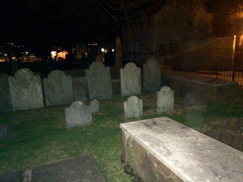 A dimly lit graveyard with various old gravestones and a stone bench, surrounded by trees and a fence.