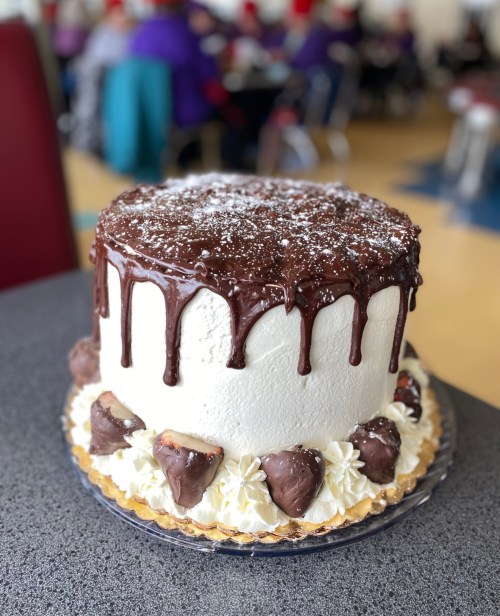 A large, round cake with white frosting, chocolate ganache drips, and chocolate-covered strawberries on the bottom.