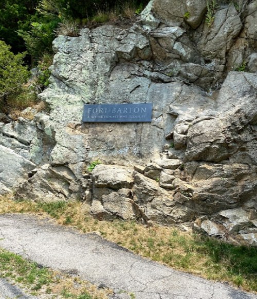 Sign for Fort Barton mounted on a rocky hillside, with a paved path leading nearby. Green foliage surrounds the area.
