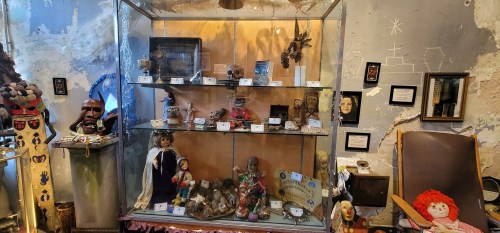 A glass display case filled with various dolls, figurines, and artifacts, set against a textured wall.