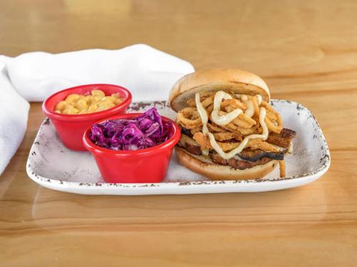 A plate with a sandwich topped with fried onions, served with red cabbage slaw and macaroni and cheese in red bowls.