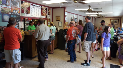 A busy restaurant interior with a line of customers waiting at the counter, looking at the menu on the wall.