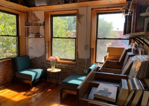 Cozy reading nook with two chairs, a small table, and shelves of books, surrounded by bright windows and greenery.