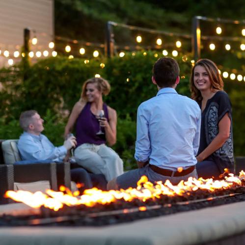 A group of friends enjoying an outdoor gathering by a fire feature, with string lights in the background.
