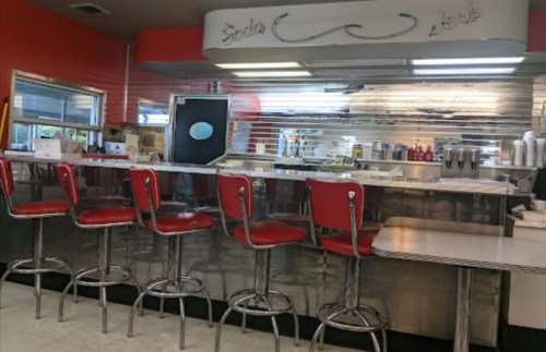 A retro diner interior featuring red bar stools, a chrome counter, and a bright red wall.