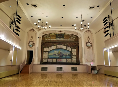 A spacious, vintage hall with a wooden floor, ornate stage backdrop, and decorative lighting fixtures.