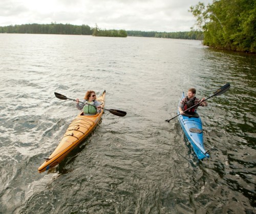 Two people kayaking on a calm lake, one in a yellow kayak and the other in a blue kayak, surrounded by trees.