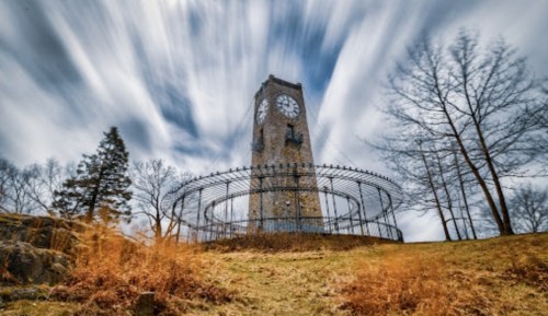 A tall clock tower surrounded by a wrought iron fence, set against a dramatic sky with moving clouds.
