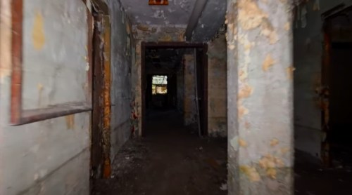 A dimly lit, abandoned hallway with peeling walls and a view of a darkened room at the end.
