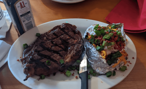 A grilled steak with grill marks, served alongside a loaded baked potato topped with cheese, bacon, and green onions.