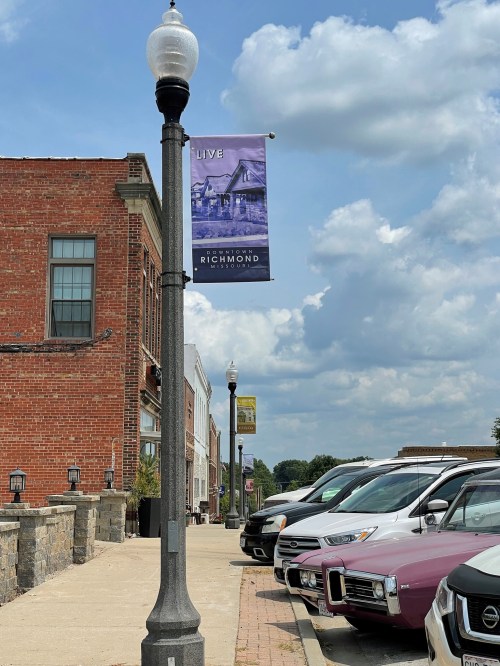 Street view featuring a lamp post with a banner, parked cars, and brick buildings under a partly cloudy sky.