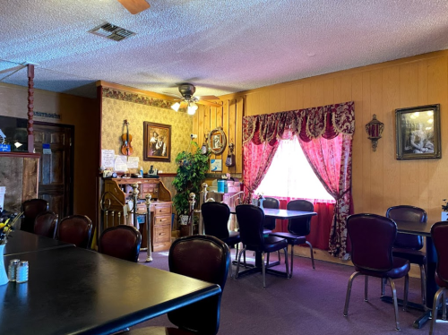 Cozy dining area with dark tables, chairs, and warm decor, featuring a window with red curtains and vintage wall art.