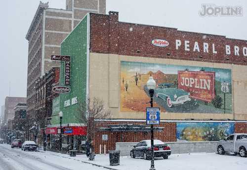 Snow falls over a street scene featuring a vintage mural and buildings in Joplin, Missouri. Cars are parked along the road.