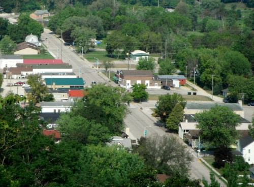 Aerial view of a small town with green trees, buildings, and a quiet street lined with shops and homes.