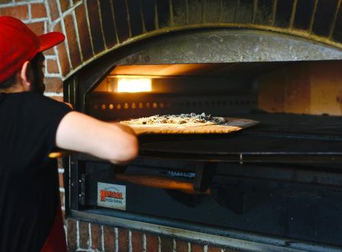 A person in a red hat places a pizza into a brick oven with a warm glow inside.