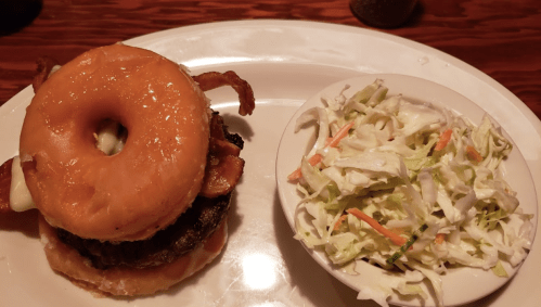 A burger with a glazed donut bun, bacon, and a side of coleslaw on a white plate.