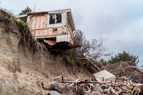 A partially collapsed house teeters on a cliff's edge, surrounded by fallen trees and debris.