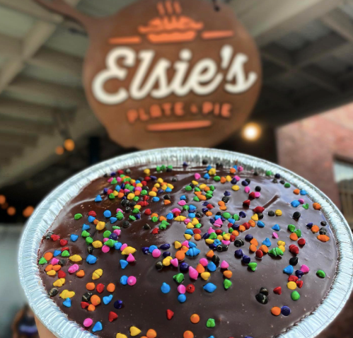 A chocolate pie topped with colorful sprinkles, held in front of a sign for "Elsie's Plate & Pie."
