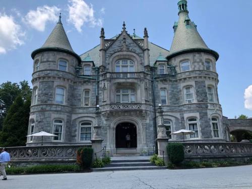 A grand stone mansion with turrets and a large entrance, surrounded by greenery and blue skies.