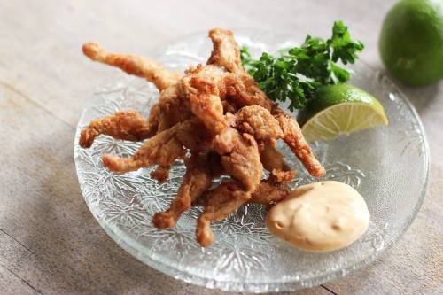 Fried frog legs on a glass plate, garnished with lime, parsley, and a side of dipping sauce.