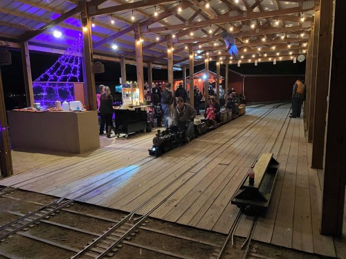 A miniature train runs on tracks under a lit pavilion, with people enjoying food and festivities in the background.