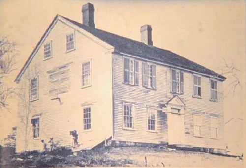 Historic two-story house with a slanted roof, multiple windows, and a front door, set in a rural landscape.