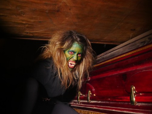 A woman with green face paint and wild hair emerges from a coffin, showcasing a fierce expression in a dark setting.