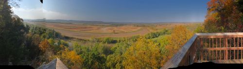 A panoramic view of rolling hills and fields, framed by colorful autumn foliage and a clear blue sky.