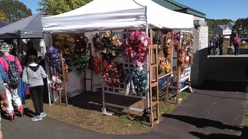 A market stall displaying colorful wreaths and floral arrangements, with people browsing nearby.