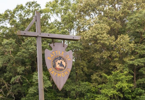 Sign for the Battle of Raymond, featuring a shield design, set against a backdrop of green trees.