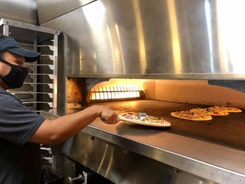 A person wearing a mask removes a pizza from a commercial oven, with more pizzas baking inside.