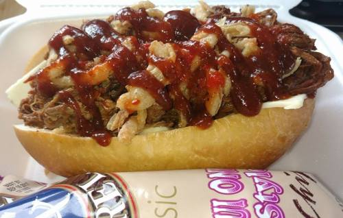 A loaded sandwich with pulled meat, crispy onions, barbecue sauce, and a side of chips on a white plate.