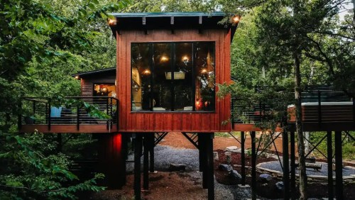 A modern wooden cabin on stilts surrounded by trees, featuring large windows and warm lighting.