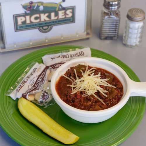 A bowl of chili topped with cheese, served with a pickle and oyster crackers on a green plate, with salt and pepper shakers in the background.