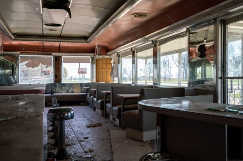 Abandoned diner interior with empty booths, broken windows, and debris scattered on the floor.