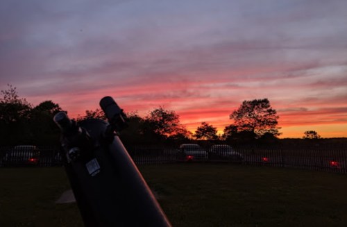 A telescope in the foreground with a vibrant sunset and colorful clouds in the background.