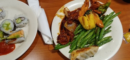A plate of green beans, orange chicken, and dumplings next to a plate of sushi rolls on a wooden table.