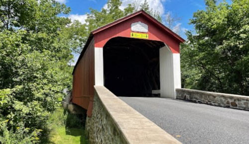 A red covered bridge with a stone approach, surrounded by green trees and a blue sky.
