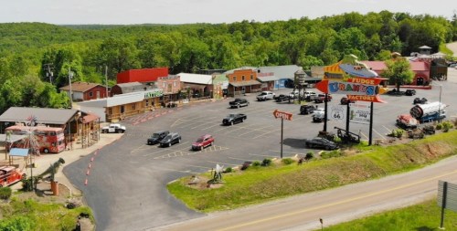 Aerial view of a small roadside shopping area with colorful buildings, parked cars, and green hills in the background.