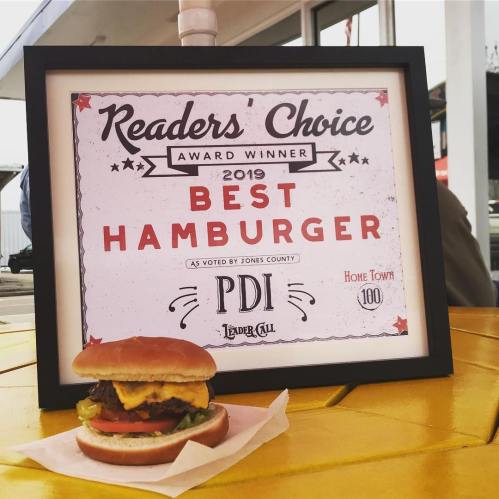 A framed award for "Best Hamburger" with a burger on a table, highlighting the 2019 Readers' Choice win.